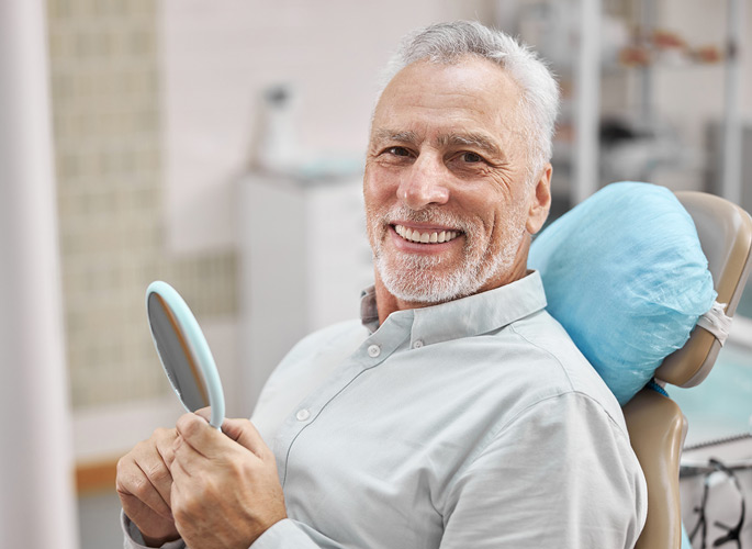 Older man smiling in dental chair holding a mirror
